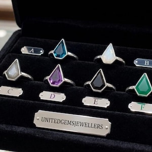 May include: A selection of gemstone rings displayed in a black case. The rings showcase various geometric-cut gemstones in colours including teal, purple, and green, set in silver bands. Small metal tags with letters accompany each ring.