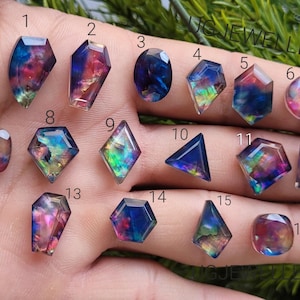 Può includere: Assortimento di 16 pietre preziose cabochon multicolori iridescenti. Le pietre preziose sono disposte in file su una mano, con i numeri da 1 a 16 che etichettano ogni pietra.