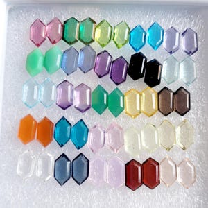 Op de afbeelding: Assortiment van 24 gefacetteerde edelsteen cabochons in verschillende kleuren, waaronder groen, blauw, paars, roze, oranje en bruin. De cabochons zijn gerangschikt in rijen op een witte achtergrond.