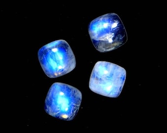 May include: Four iridescent blue moonstone cabochons, each with a square shape and a smooth, polished surface. The stones are arranged in a scattered pattern on a black background.