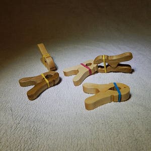 Puede incluir: Cuatro pinzas de madera con gomas elásticas. Las pinzas son de varios colores y están dispuestas sobre una superficie marrón claro.