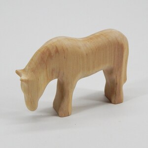 May include: A wooden horse figurine with a light brown color. The horse is standing with its head lowered as if it is grazing.