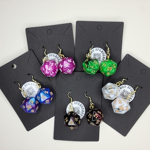 Puede incluir: Pendientes con dados multicolores de múltiples caras en varios colores, incluyendo morado, verde, azul, negro y blanco. Cada pendiente tiene un gancho dorado. Los dados se muestran en tarjetas negras.