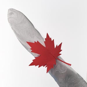 May include: A red maple leaf napkin ring adorns a silver napkin. The leaf is made of paper and has a detailed, jagged edge. The napkin has a subtle floral pattern. This decorative item is suitable for fall or Canadian-themed events.