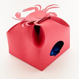 Peut inclure: Une boîte en papier rouge avec une découpe de crabe sur le dessus. La boîte a un petit trou sur le côté, révélant des bonbons bleus à l'intérieur.