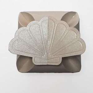Puede incluir: Una caja de cartón beige con un diseño de concha de mar de purpurina plateada en la parte superior. La caja está doblada y lista para ser ensamblada.