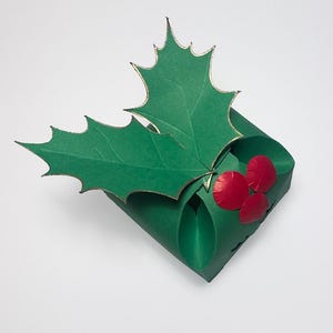 May include: A green paper gift box with a holly leaf design and red berries. The box is folded and ready to be filled with a small gift.