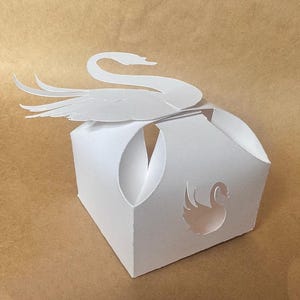May include: A white paper box with a swan design. The box is folded into a cube shape with a swan cutout on the front. The swan is also the lid of the box.