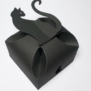 Puede incluir: Una caja de regalo de papel negro con una silueta de gato negro recortada en la parte superior. El gato está sentado con la cola enroscada alrededor de su cuerpo.