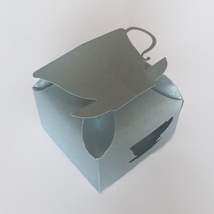 May include: A light blue paper favor box shaped like a teacup with a cutout of a teacup on the side.