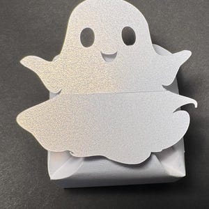 May include: A white ghost-shaped box with a smiling face and two eyes. The box is made of paper and has a glittery finish.