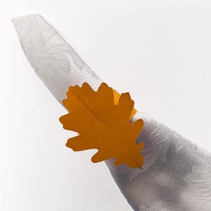 May include: A decorative napkin ring shaped like a fall leaf in shades of orange and yellow. The leaf-shaped ring is wrapped around a folded white napkin with a subtle floral pattern, creating a seasonal table setting.