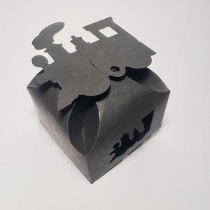 Könnte beinhalten: Dunkelgraue Geschenkbox aus Papier mit einer Zugsilhouette oben und einem Zug-Ausschnitt an der Seite. Die Box ist quadratisch, mit gefalteten Kanten und einem dekorativen Zug-Ausschnitt.