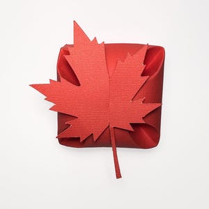 Puede incluir: Una hoja de arce de papel rojo se coloca encima de una caja de regalo cuadrada roja. La hoja está recortada con bordes detallados. La caja de regalo está envuelta en papel rojo con esquinas dobladas.