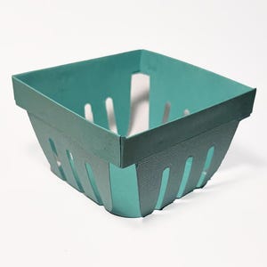 May include: A teal-colored, square berry basket made of cardboard. The basket has a rectangular shape with vertical slots on the sides for ventilation. The top edge is reinforced with a darker teal band.