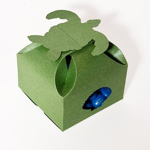 Peut inclure: Boîte à dragées verte en forme de tortue avec une découpe pour les friandises. La boîte est en carton et a une finition texturée.