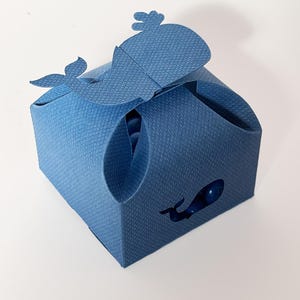 Op de afbeelding: Een blauwe walvisvormige cadeaudoos met een uitgesneden walvisontwerp aan de voorkant. De doos is gemaakt van karton en heeft een textuur.