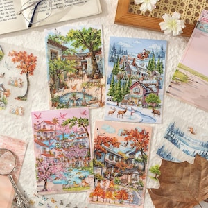 May include: Four decorative paper cards with scenic illustrations. One depicts a winter village with Santa's sleigh, another a spring scene with cherry blossoms, and two others show autumn and summer landscapes. A magnifying glass and a book are also visible.