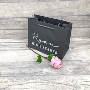 May include: A grey gift bag with white text that reads "Ryan Ring Bearer". A pink flower is lying on a grey wooden surface.