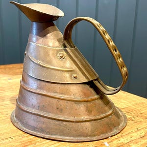 May include: An antique copper jug with a distinctive, layered design. It has a curved handle and a spout. The metal has a warm, aged patina, indicating it's vintage. The handle is decorated with small holes. A decorative piece.