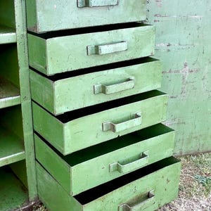 K&ouml;nnte beinhalten: Ein gr&uuml;ner Vintage-Metallschrank mit f&uuml;nf Schubladen, jede mit einem rechteckigen Griff. Der Schrank hat ein verwittertes Aussehen, das auf Alter und Nutzung hindeutet. Die offene T&uuml;r zeigt Regale. Der Schrank steht drau&szlig;en auf Gras.