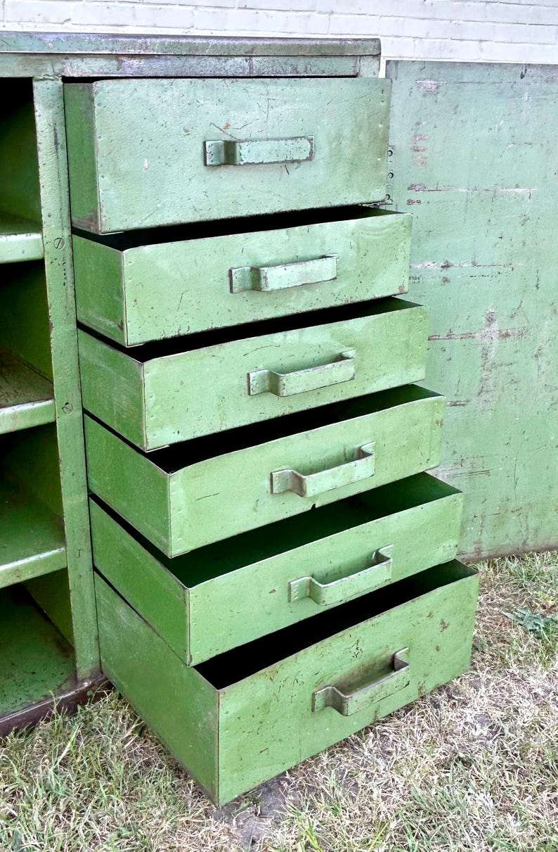 K&ouml;nnte beinhalten: Ein gr&uuml;ner Vintage-Metallschrank mit f&uuml;nf Schubladen, jede mit einem rechteckigen Griff. Der Schrank hat ein verwittertes Aussehen, das auf Alter und Nutzung hindeutet. Die offene T&uuml;r zeigt Regale. Der Schrank steht drau&szlig;en auf Gras.