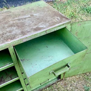 K&ouml;nnte beinhalten: Ein gr&uuml;ner Metallschrank im Vintage-Stil mit einer offenen Schublade. Der Schrank hat eine rechteckige Form mit einem leicht verwitterten Aussehen. Die Schublade hat einen Metallgriff. Der Schrank steht im Freien auf Gras.