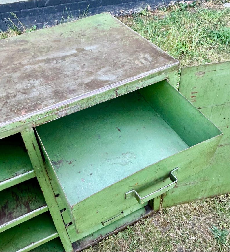 K&ouml;nnte beinhalten: Ein gr&uuml;ner Metallschrank im Vintage-Stil mit einer offenen Schublade. Der Schrank hat eine rechteckige Form mit einem leicht verwitterten Aussehen. Die Schublade hat einen Metallgriff. Der Schrank steht im Freien auf Gras.