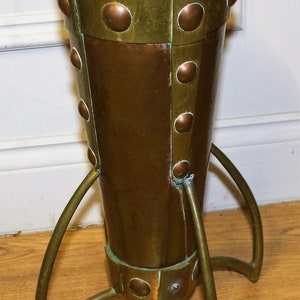May include: A copper and brass umbrella stand with a conical shape and a three-legged base. The stand has a hammered finish and decorative rivets.