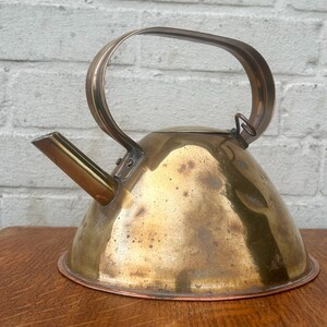 May include: A vintage brass kettle with a copper base and a curved handle. The kettle has a spout and a lid. The brass has a warm, aged patina. The kettle is likely used for boiling water on a stovetop.