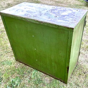 K&ouml;nnte beinhalten: Ein verwitterter, gr&uuml;ner Metallschrank mit rechteckiger Form. Der Schrank hat eine leicht abgeschr&auml;gte Oberseite mit einer kreisf&ouml;rmigen Vertiefung. Die Farbe weist Gebrauchsspuren auf, was ihm ein Vintage-Aussehen verleiht. Der Schrank steht auf einer grasbewachsenen Oberfl&auml;che.