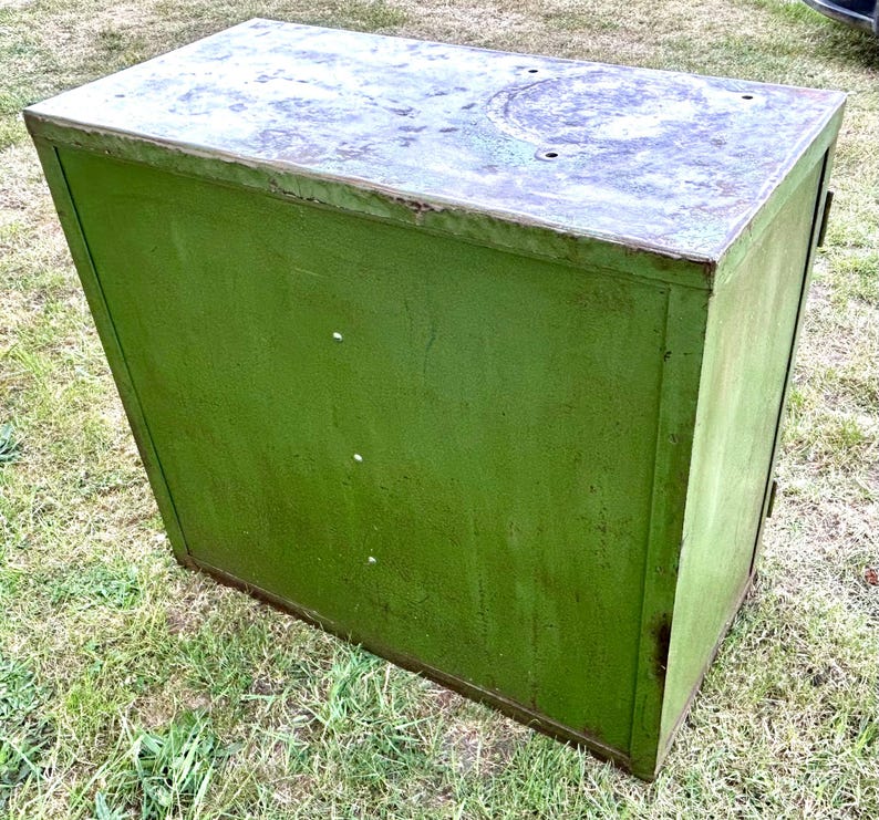 K&ouml;nnte beinhalten: Ein verwitterter, gr&uuml;ner Metallschrank mit rechteckiger Form. Der Schrank hat eine leicht abgeschr&auml;gte Oberseite mit einer kreisf&ouml;rmigen Vertiefung. Die Farbe weist Gebrauchsspuren auf, was ihm ein Vintage-Aussehen verleiht. Der Schrank steht auf einer grasbewachsenen Oberfl&auml;che.