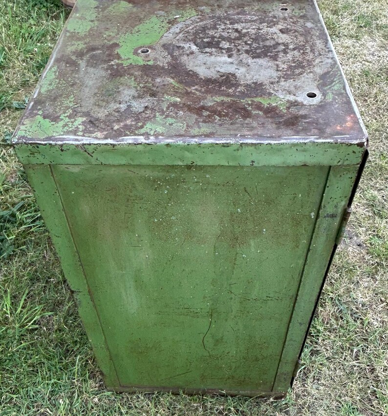K&ouml;nnte beinhalten: Ein verwitterter, gr&uuml;ner Metallschrank mit rechteckiger Form. Die Oberseite weist Rost- und Abnutzungserscheinungen auf, mit mehreren kleinen L&ouml;chern. Die Seiten des Schranks sind einfarbig, verblasst gr&uuml;n, was auf Alter und Nutzung hindeutet. Der Schrank steht im Freien auf Gras.