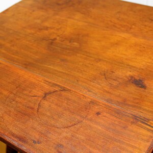 May include: A close-up of a wooden table top with a light brown finish. The surface shows signs of wear and tear, including several circular marks.