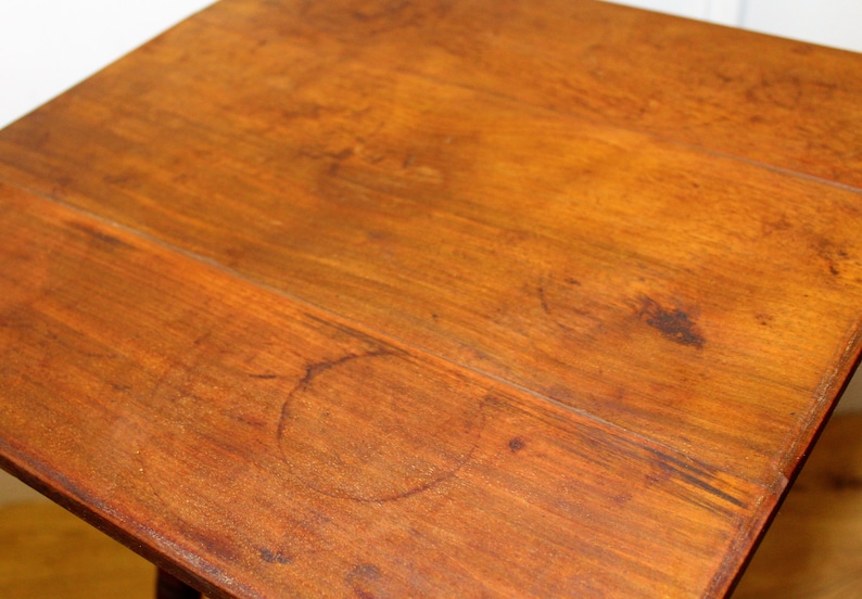 May include: A close-up of a wooden table top with a light brown finish. The surface shows signs of wear and tear, including several circular marks.
