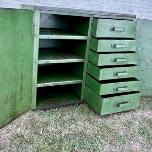 K&ouml;nnte beinhalten: Ein antiker gr&uuml;ner Metallschrank mit offenen T&uuml;ren und Schubladen. Der Schrank hat Regale und sechs Auszugsschubladen auf der rechten Seite. Der Schrank ist verwittert und weist Alterserscheinungen auf.