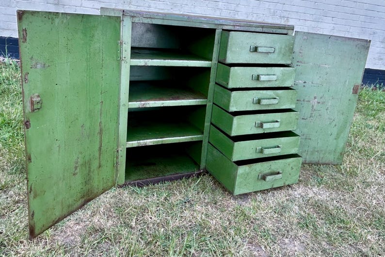 K&ouml;nnte beinhalten: Ein antiker gr&uuml;ner Metallschrank mit offenen T&uuml;ren und Schubladen. Der Schrank hat Regale und sechs Auszugsschubladen auf der rechten Seite. Der Schrank ist verwittert und weist Alterserscheinungen auf.