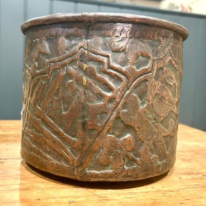 May include: A copper-toned, cylindrical vessel featuring detailed, carved floral and foliate patterns. The relief work includes leaves, vines, and possibly birds, covering the entire surface. The container has a slightly flared rim.