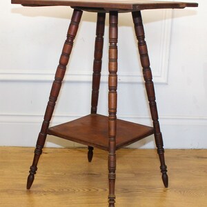 May include: A dark brown wooden table with a square top and three legs. The legs are turned and have a decorative design. The table has a lower shelf.