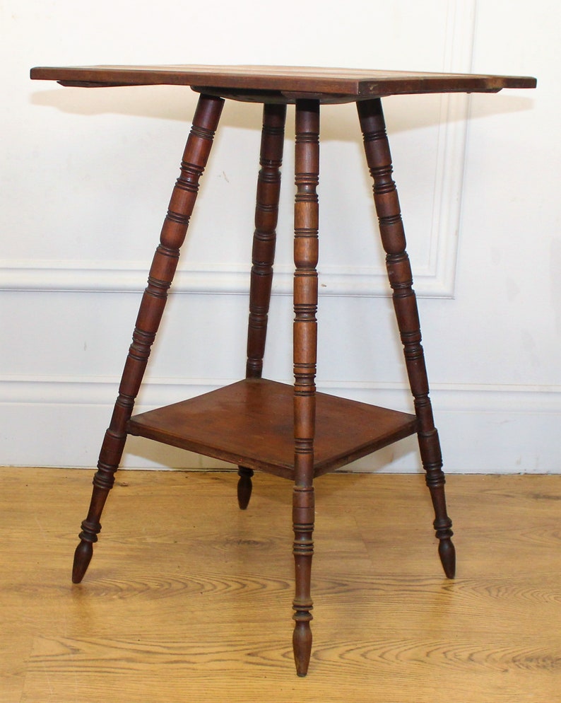May include: A dark brown wooden table with a square top and three legs. The legs are turned and have a decorative design. The table has a lower shelf.