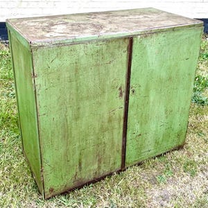 K&ouml;nnte beinhalten: Ein verwitterter, gr&uuml;ner Metallschrank mit zwei T&uuml;ren. Der Schrank hat eine rechteckige Form und ein leicht abgenutztes Aussehen, was auf Alter und Nutzung hindeutet. Die T&uuml;ren treffen sich in der Mitte mit einer vertikalen Naht. Der Schrank steht drau&szlig;en auf Gras.