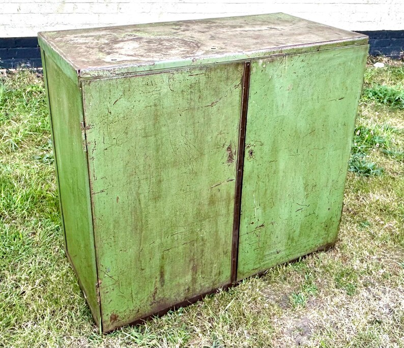 K&ouml;nnte beinhalten: Ein verwitterter, gr&uuml;ner Metallschrank mit zwei T&uuml;ren. Der Schrank hat eine rechteckige Form und ein leicht abgenutztes Aussehen, was auf Alter und Nutzung hindeutet. Die T&uuml;ren treffen sich in der Mitte mit einer vertikalen Naht. Der Schrank steht drau&szlig;en auf Gras.