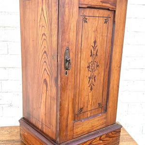 May include: A tall, rectangular wooden cabinet with a natural light brown wood grain finish. The front door features a dark brown decorative stencil pattern and a metal drop handle with a keyhole. The cabinet sits on a thick, raised base.