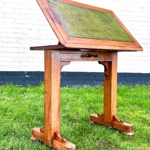 May include: An antique wooden writing desk with a green leather writing surface. The desk has a slanted top, a small drawer, and is supported by two sturdy legs. The desk is made of wood and is set on a grassy lawn.