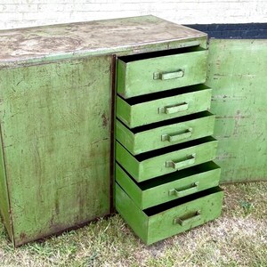K&ouml;nnte beinhalten: Ein gr&uuml;ner Vintage-Metallschrank mit sechs Schubladen und einer offenen Seitent&uuml;r. Der Schrank hat eine abgenutzte Oberfl&auml;che, die Gebrauchsspuren aufweist. Die Schubladen haben Metallgriffe. Der Schrank steht auf Gras.