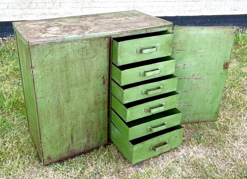 K&ouml;nnte beinhalten: Ein gr&uuml;ner Vintage-Metallschrank mit sechs Schubladen und einer offenen Seitent&uuml;r. Der Schrank hat eine abgenutzte Oberfl&auml;che, die Gebrauchsspuren aufweist. Die Schubladen haben Metallgriffe. Der Schrank steht auf Gras.