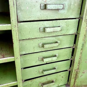 K&ouml;nnte beinhalten: Ein gr&uuml;ner Vintage-Metallschrank mit f&uuml;nf Schubladen und offenen Regalen. Der Schrank hat eine abgenutzte Oberfl&auml;che, die Gebrauchsspuren und Alter aufweist. Die Schubladen haben Metallgriffe.