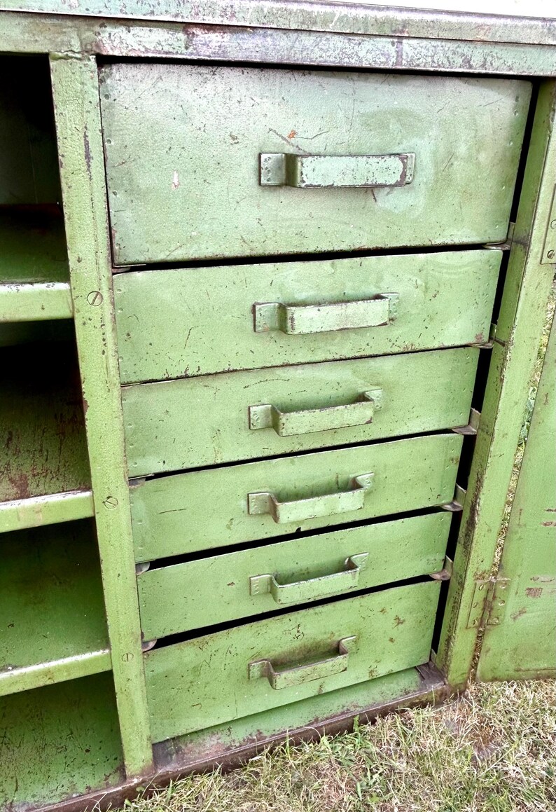 K&ouml;nnte beinhalten: Ein gr&uuml;ner Vintage-Metallschrank mit f&uuml;nf Schubladen und offenen Regalen. Der Schrank hat eine abgenutzte Oberfl&auml;che, die Gebrauchsspuren und Alter aufweist. Die Schubladen haben Metallgriffe.