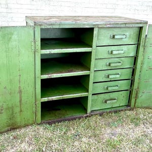 K&ouml;nnte beinhalten: Ein gr&uuml;ner Vintage-Metallschrank mit f&uuml;nf Schubladen und mehreren Regalen. Die Schrankt&uuml;ren sind ge&ouml;ffnet und geben den Blick ins Innere frei. Der Schrank steht auf einer grasbewachsenen Oberfl&auml;che.
