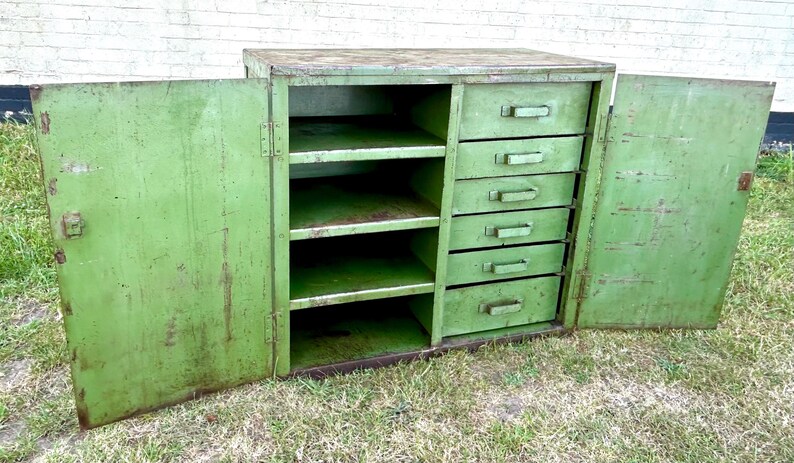 K&ouml;nnte beinhalten: Ein gr&uuml;ner Vintage-Metallschrank mit f&uuml;nf Schubladen und mehreren Regalen. Die Schrankt&uuml;ren sind ge&ouml;ffnet und geben den Blick ins Innere frei. Der Schrank steht auf einer grasbewachsenen Oberfl&auml;che.